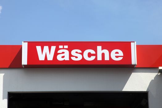 Sign in German above the entrance to an automatic car wash saying Wasche on a red background against a clear blue sky