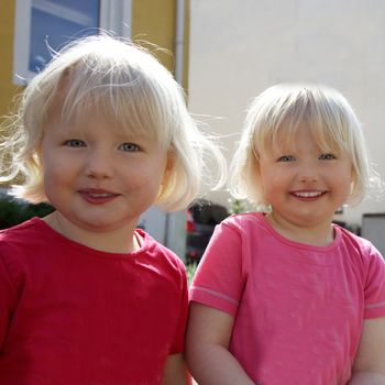 Pair of cute little blond identical twins with mischievous expressions standing looking at the camera, closeup upper body portrait