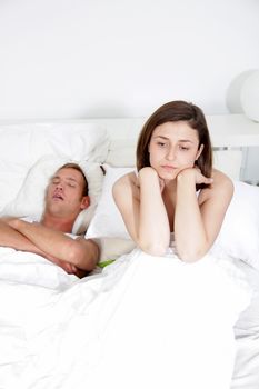 Man lying on his back in bed snoring loudly while his wife, unable to sleep, sits up with a resigned angry expression