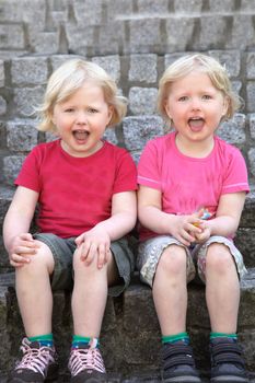 Adorable happy identical blond twins sitting close together against a stone wall with their mouths open