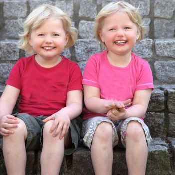 Laughing cute identical blond twins sitting against a stone wall looking at the camera with charming smiles
