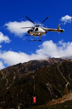 Small helicopter with transport cargo bucket flying in blue sky over mountains of Southern Alps New Zealand to supply remote site