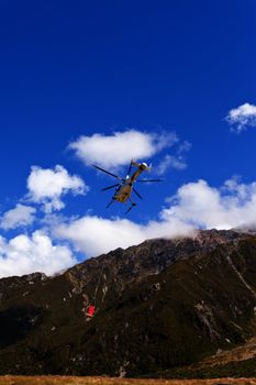 Small helicopter with transport cargo bucket flying in blue sky over mountains of Southern Alps New Zealand to supply remote site