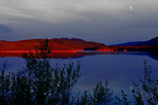 Red glow of evening sun on boreal forest taiga surrounding calm water surface of Lake Twin Lake near Carmacks Yukon Territory Canada