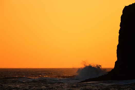 Beautiful marine sunset with a sky suffused by orange light and a wave crashing onto the base of a high cliff