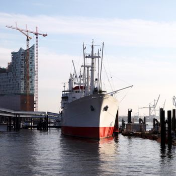 Commercial ship or trawler docked alongside a jetty in a marine harbour with a construction site on a highrise building and skywalk behind it