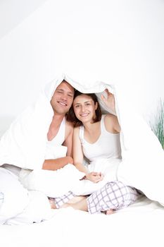 Smiling young couple sitting on their bed under the duvet which they have pulled over their heads