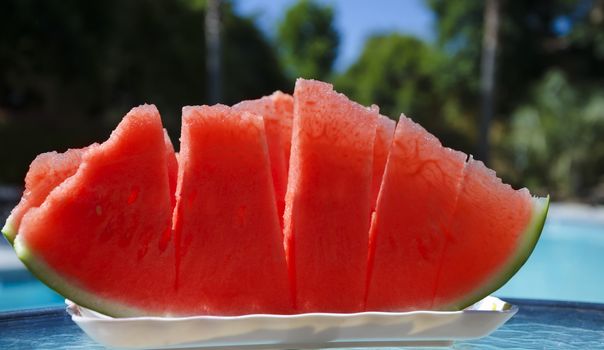 Few peaces of watermelon in white plate on table by the swimming pool