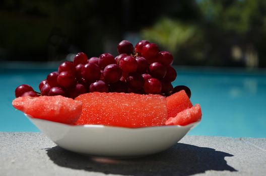 Grapes and watermelon in white plate on table by the swimming pool