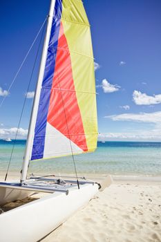 Small sailing catamaran with a colourful striped sail beached on a sandy tropical beach