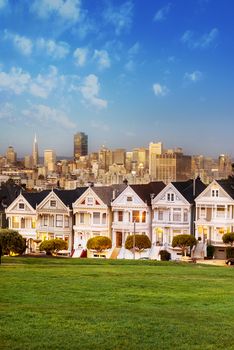 The Painted Ladies of San Francisco, California sit glowing amid the backdrop of a sunset and skyscrapers. 