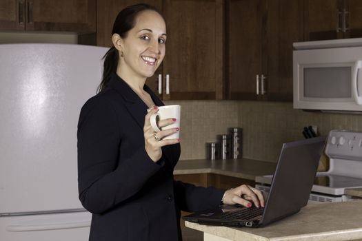 Business woman working on a laptop computer and drinking coffer in her kitchen.