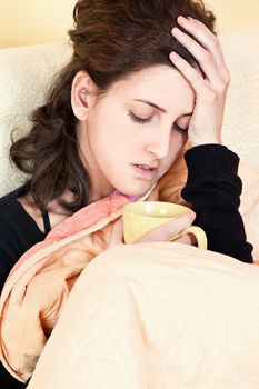 A young sick woman with a cup of tea covered with blanket at home, holding her head in pain