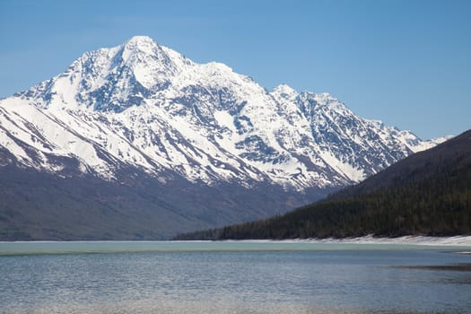 A large snow covered mountain rises above clear blue water