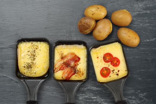 Photo of a three Raclette trays with melted cheese, herbs, cherry tomato, bacon, onion and potatoes on rough slate. Plenty of space for text.