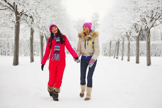 two women walk by winter alley snow trees on background