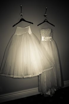 Two beautiful filmy wedding gowns with graceful full skirts hanging on clothes hangers on a wall in a darkened room