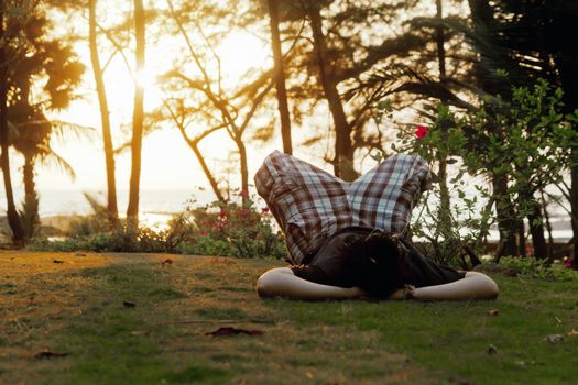 Horizontal color landscape of man laying on garden lawn, arms acting as support for head, point of view at his head, knees folded up, sunset shinning through the trees, location of shot Manori, Bombay, India