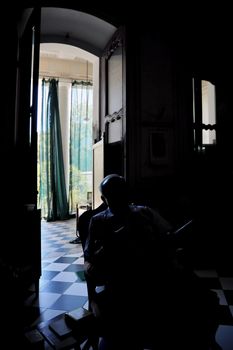 Vertical portrait of a seated male man gentleman near coffee table of books backlit from a doorway of an old building with architectural interest. Shot location was the Ashram at Pondicherry, Tamil Nadu India