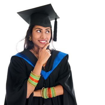 Happy Indian graduate student in graduation gown and cap thinking and smiling. Portrait of beautiful Asian female model standing isolated on white background.