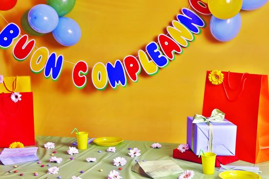 Birthday party, a green table with gifts, flowers and candy particular scattered everywhere, on the yellow background colorful balloons and streamers Wishing happy birthday.