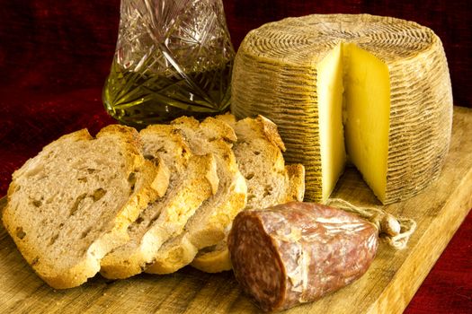 Mediterranean food: sheep cheese, salami, bread with walnuts and olive oil on a wooden cutting board on a dark red background.