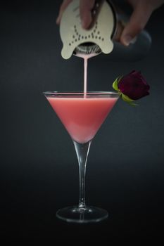 Hand with shaker pouring a pink cocktail made with vodka, yogurt liquor and rose syrup decorated with rosebud on dark background.