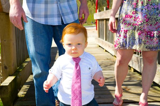 A baby wearing a red or pink necktie walks across a bridge with his parents behind him.