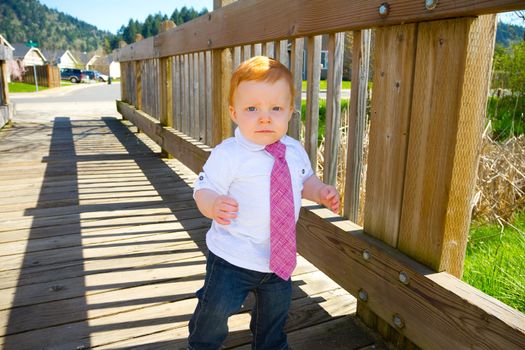 A one year old baby boy walks across a bridge wearing nice clothes and a necktie.