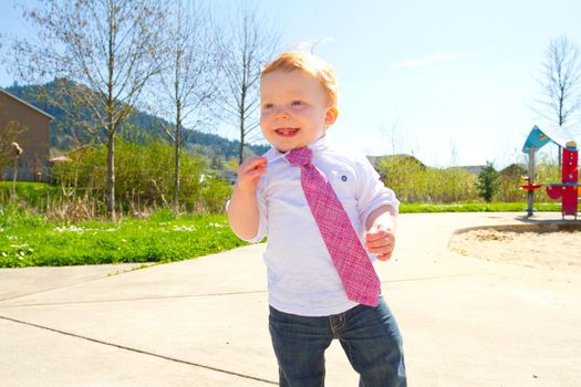 A baby boy plays at the park wearing his Sunday's best clothes including a tie around his neck.