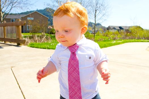 A baby boy plays at the park wearing his Sunday's best clothes including a tie around his neck.