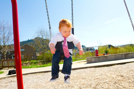 A baby boy plays on a swing set at the park wearing nice clothing.