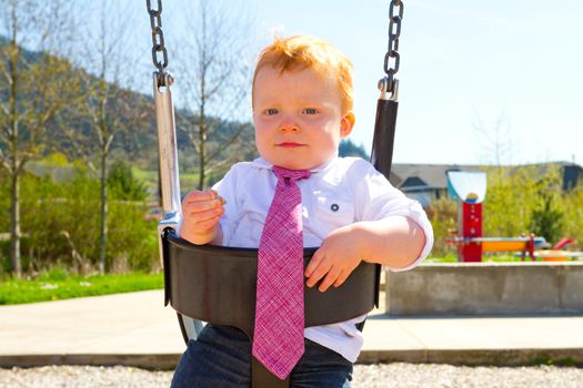 A baby boy plays on a swing set at the park wearing nice clothing.