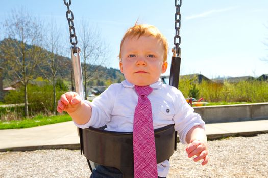 A baby boy plays on a swing set at the park wearing nice clothing.