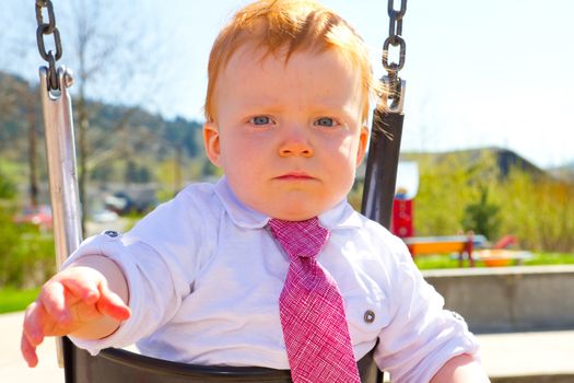 A baby boy plays on a swing set at the park wearing nice clothing.