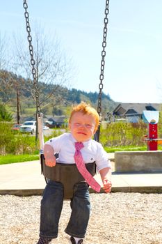 A baby boy sits on a swing set while upset and crying wearing nice clothes.