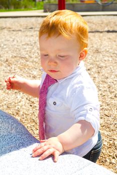 A caucasian baby boy plays at the park wearing a white shirt and a tie.