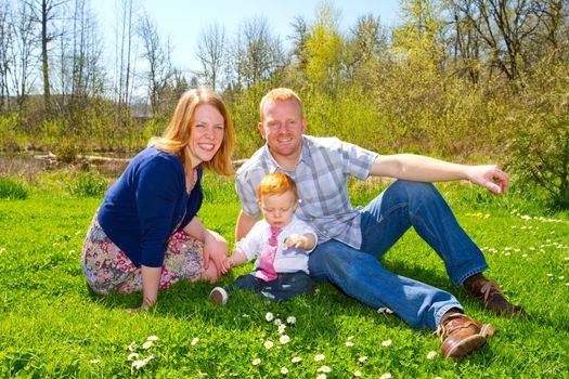 A mom and dad with their one year old baby boy at a nice park wearing trendy clothes.