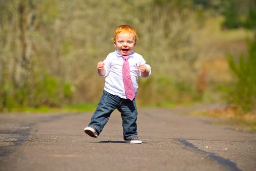 A one year old boy taking some of his first steps outdoors on a path with selective focus while wearing a nice shirt and a necktie.