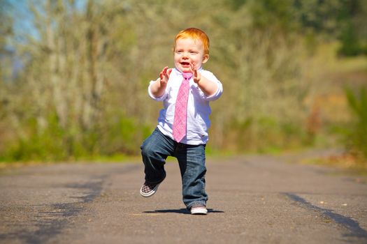 A one year old boy taking some of his first steps outdoors on a path with selective focus while wearing a nice shirt and a necktie.