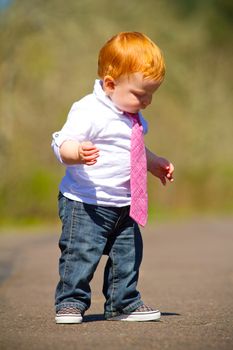 A one year old boy taking some of his first steps outdoors on a path with selective focus while wearing a nice shirt and a necktie.