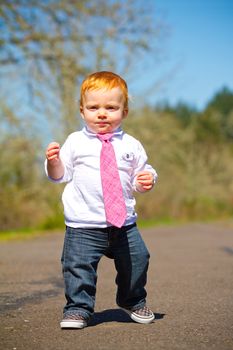 A one year old boy taking some of his first steps outdoors on a path with selective focus while wearing a nice shirt and a necktie.