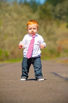 A one year old boy taking some of his first steps outdoors on a path with selective focus while wearing a nice shirt and a necktie.