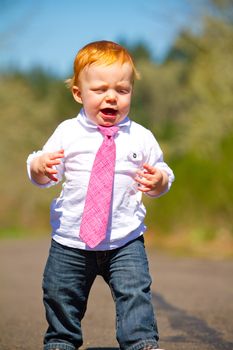 A one year old boy taking some of his first steps outdoors on a path with selective focus while wearing a nice shirt and a necktie.