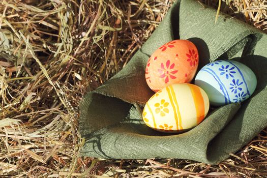 Three colored Easter eggs and green textile on hay at sunny day