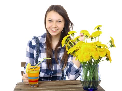 One teenage girl, caucasian, 16, sitting at a table with a glass of drink. A girl dressed in a plaid shirt. Girl smiles. On table is a blue vase, with yellow wildflowers, a bouquet of dandelions.