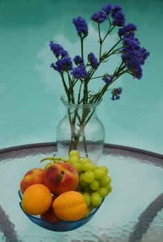 Vase with blue flowers and apricots, grape and peaches in plate are on the glass table by the swimming pool