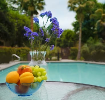 Vase with blue flowers and apricots, grape and peaches in plate are on the glass table by the swimming pool