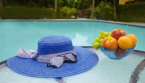 Woman's hat, sunglasses and grape with peaches and apricots in glass plate are on table by the swimming pool