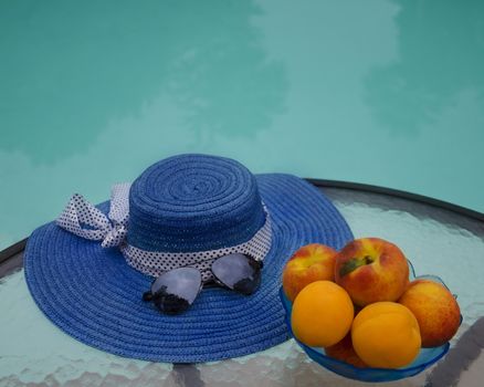 Woman's hat, sunglasses and peaches in glass plate are on table by the swimming pool